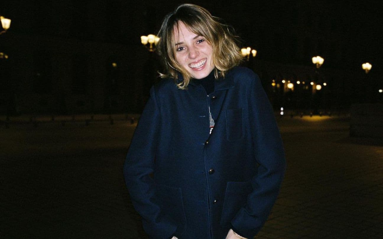 Maya Hawke sorri para foto, com as mãos no bolso