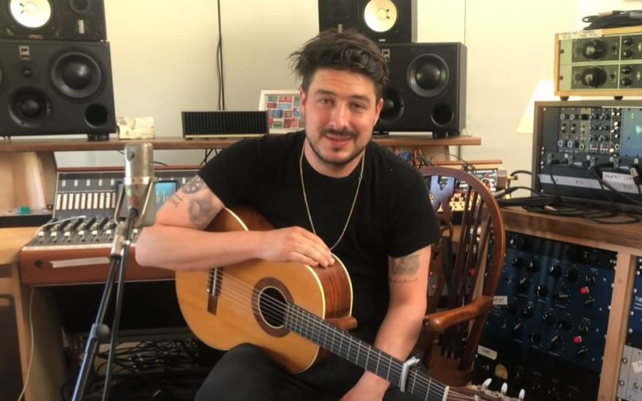 Marcus Mumford segura um violão, sentado em uma cadeira de estúdio