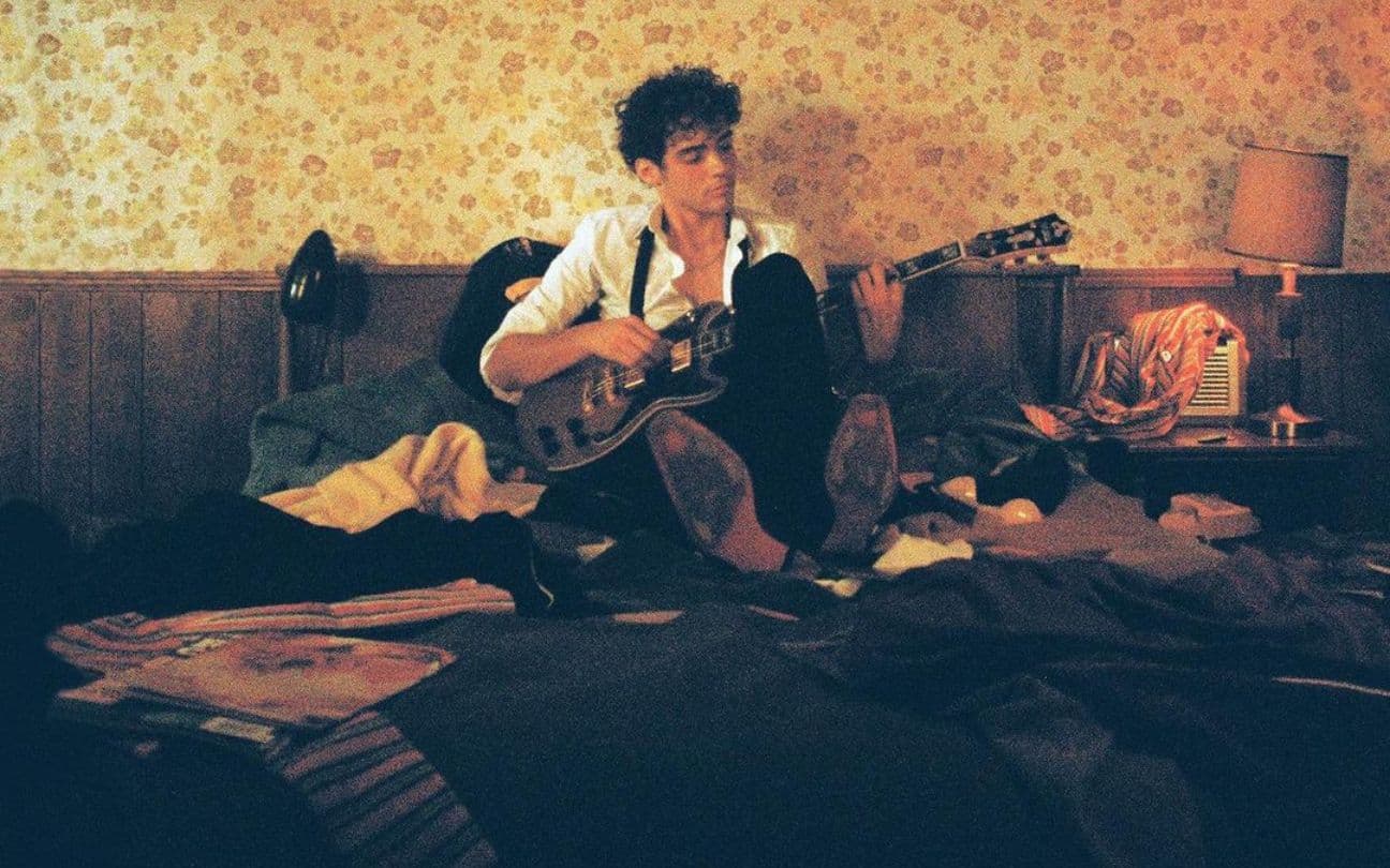 Joshua Basset segura violão, sentada em cama de hotel